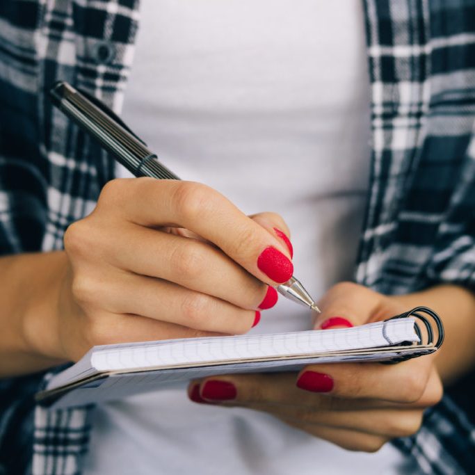 Woman in plaid shirt and a red manicure pen writing in a notebook